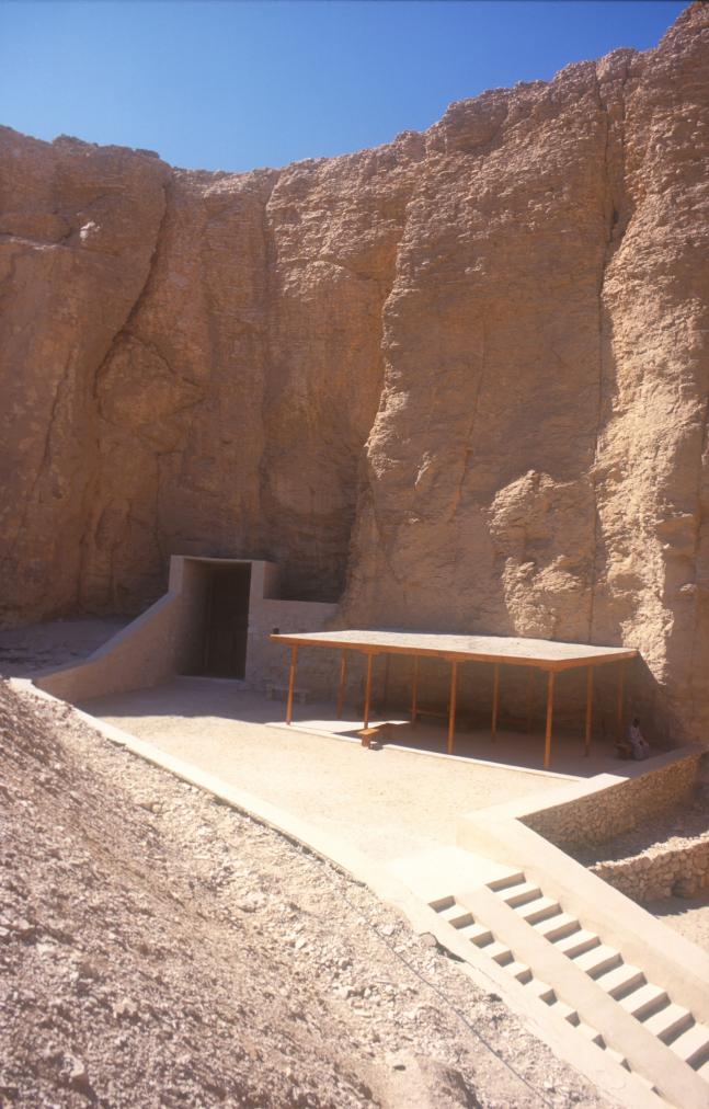 General view of the tomb entrance at base of cliff with modern covering, adjacent shelter, and modern approach path.